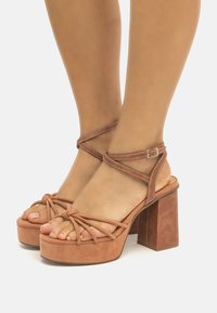 Sandalias de plataforma de ante marrón con un grueso tacón cuadrado, tira de tobillo con diseño entrelazado y punta con nudo. Textura suave con un acabado mate.
