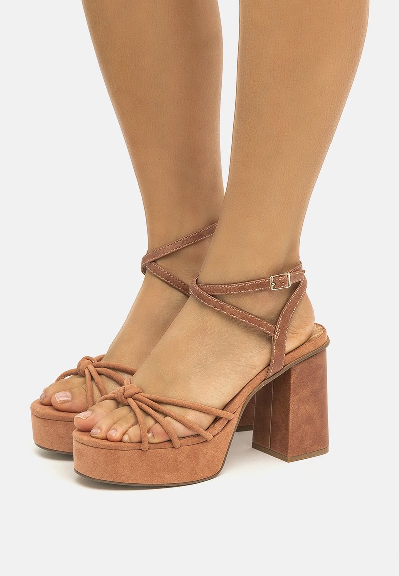 Sandalias de plataforma de ante marrón con un grueso tacón cuadrado, tira de tobillo con diseño entrelazado y punta con nudo. Textura suave con un acabado mate.