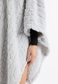 Szary polarowy poncho z fakturowanym wzorem i pluszowym wykończeniem, z podwiniętym brzegiem. Widoczna jest ręka trzymająca krawędź materiału.