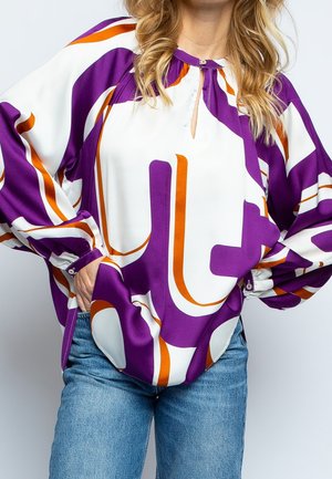 Mujer que lleva una blusa suelta de manga larga con un patrón abstracto en tonos morado, blanco y naranja, metida dentro de vaqueros azules, con las manos en los bolsillos.