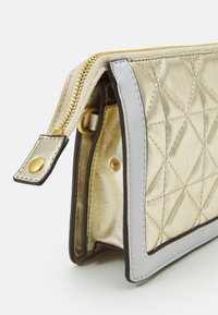 Bolso acolchado dorado con borde blanco, cremallera dorada parcialmente abierta, mostrando un botón de presión lateral y detalle de anillo dorado.