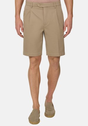 Mężczyzna ubrany w beżowe, dopasowane spodenki i pasujące wsuwane buty, stojący na tle jednolitego tła.