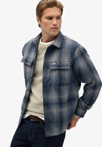 Camicia a quadri blu in flanella con chiusura a bottoni, due tasche sul petto e vestibilità comoda, indossata sopra un felpa di colore chiaro.