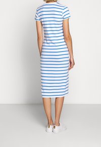 Donna che indossa un vestito midi bianco con strisce orizzontali blu e sneakers bianche, in piedi di fronte a un muro bianco semplice.