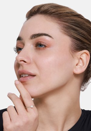 Anello in argento sterling con una piccola perla rotonda, indossato su una mano vicino al viso, con tono di pelle uniforme e trucco naturale.