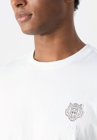 Camiseta de algodón blanca con un gráfico texturizado de un tigre en el lado izquierdo del pecho. Diseño clásico de cuello redondo con mangas cortas.