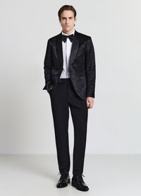 Schwarzer Smoking mit verziertem Muster, Satinrevers, weißem Hemd und schwarzer Fliege. Kombiniert mit maßgeschneiderten schwarzen Hosen und glänzenden schwarzen Schuhen.