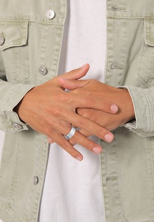 Personne portant une bague argentée texturée au majeur, mains jointes, vêtue d'une veste en jean de couleur claire et d'une chemise blanche.