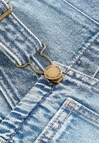 Jasnoniebieskie ogrodniczki z denimowej tkaniny o teksturalnym splocie, z metalowym zapięciem z grawerowanymi detalami i kontrastującymi białymi szwami.