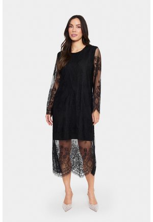 Black lace dress with long, sheer sleeves and intricate floral patterns. Sheer overlay at the hem. Paired with silver-accented shoes.