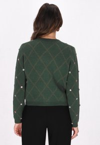 Pulover verde tricotat, cu un model în diamant, împodobit cu strasuri colorate pe mâneci. Guler rotund, finisaj texturat.
