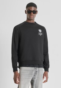 Felpa nera con maniche raglan, piccolo logo grafico bianco e collo e polsini a coste, indossata con jeans grigi. Texture liscia.