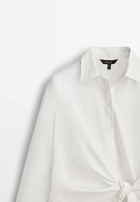 Chemise blanche à manches longues en tissu lisse avec un col classique, une fermeture à boutons sur le devant et un détail noué à la taille.
