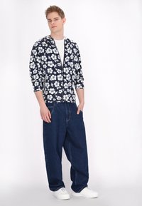 Marinblå blommig jacka med dragkedja och vitt hibiskusmönster, tillsammans med lösa mörka denimbyxor och vita sneakers.