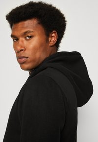 Schwarzer Kapuzenpullover mit weicher Textur und klassischer Passform, dargestellt an einem Modell mit kurzen, lockigen Haaren und im Profil.