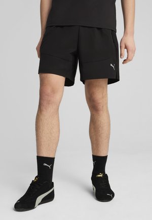 Pantalones cortos deportivos negros con una textura suave, que cuentan con dos bolsillos frontales y un pequeño logo reflectante en el lateral. Combinados con calcetines y zapatos negros.