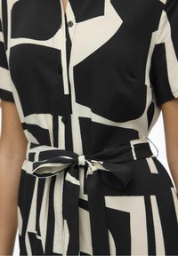 Vestido camiserо en blanco y negro con patrón geométrico, que presenta mangas cortas, cierre de botones en el frente y un cinturón atado en la cintura. Textura de tela suave.