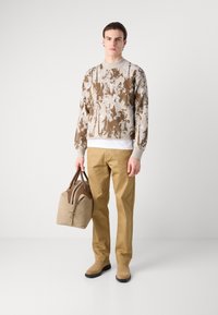 Beige sweater with a textured, abstract pattern, beige trousers, and light brown suede boots. Carrying a tan duffle bag with dark accents.