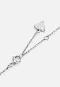 Collier en chaîne argentée avec fermoir à mousqueton et un petit pendentif triangulaire gravé du mot "GUESS" et d'un point d'interrogation.