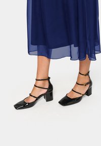 Scarpe con tacco a blocco in vernice nera, con punta quadrata e molteplici cinturini alla caviglia, con dettagli in tonalità dorata. Indossate con un vestito azzurro trasparente.
