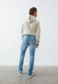 Beige hættetrøje og blå slim-fit jeans med baglommer, båret med sort-hvide sneakers på en lys grå gulv mod en hvid væg.