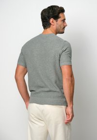 Grå kortärmad stickad tröja med rund halsringning, ribbat fåll och slät textur, bärs med ljusa byxor, visad bakifrån.