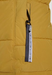 Žlutá péřová bunda s prošívaným vzorem, vybavená zipovou kapsou a textilní nálepkou s černobílým textem. Matný povrch s lesklým akcentem.