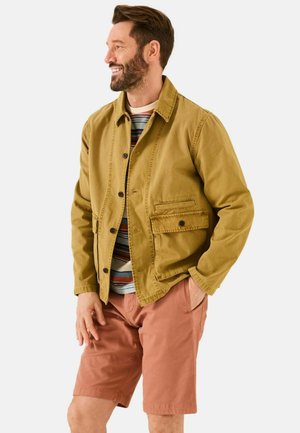 Khakifarbene Leinwandjacke mit Knöpfen und seitlicher Tasche, getragen über einem gestreiften Hemd und kombiniert mit hellrosa Shorts. Insgesamt lässiger Look.