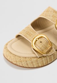 Beige flat sandal with woven straw upper, two adjustable straps, and gold-tone buckles. Soft footbed with a textured sole.
