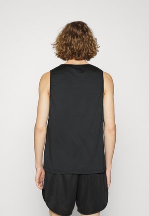 Kiiltävät kiharat hiukset omaava henkilö seisoo selin päin, pukeutuneena hihattomaan mustaan toppiin ja mustiin, teksturoituihin shortseihin yksivärisen taustan edessä.