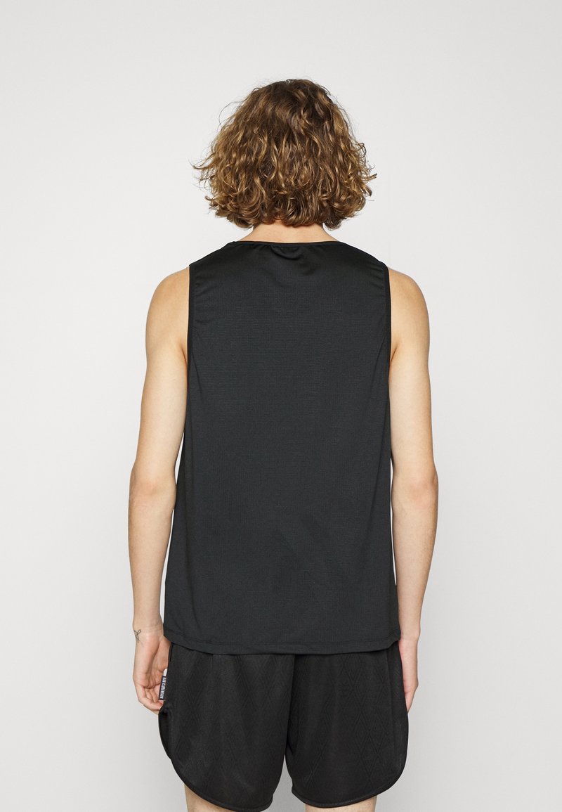 Personne aux cheveux bouclés vue de dos, portant un haut noir sans manches et un short noir texturé, devant un fond uni.