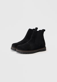 Un paio di stivali Chelsea in suede nero con pannelli elastici laterali e suole in gomma robuste su uno sfondo chiaro e uniforme.
