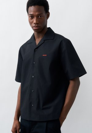 Νεαρός άνδρας που φοράει μαύρο πουκάμισο με κοντά μανίκια και κόκκινο λογότυπο "HUGO" στο στήθος, στέκεται μπροστά σε απλό λευκό φόντο.