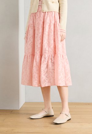 Γυναίκα με μπεζ ζακέτα και ροζ φλοράλ μίντι φούστα που στέκεται σε ξύλινο πάτωμα, φορώντας μπεζ παπούτσια Mary Jane.