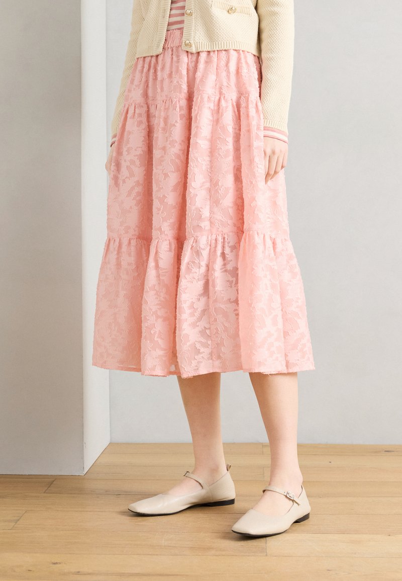 Γυναίκα με μπεζ ζακέτα και ροζ φλοράλ μίντι φούστα που στέκεται σε ξύλινο πάτωμα, φορώντας μπεζ παπούτσια Mary Jane.