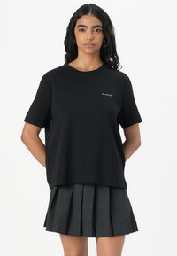 T-shirt in cotone nero con scollatura rotonda e maniche corte, caratterizzata da un piccolo logo bianco sul petto sinistro, abbinata a una gonna grigia plissettata.