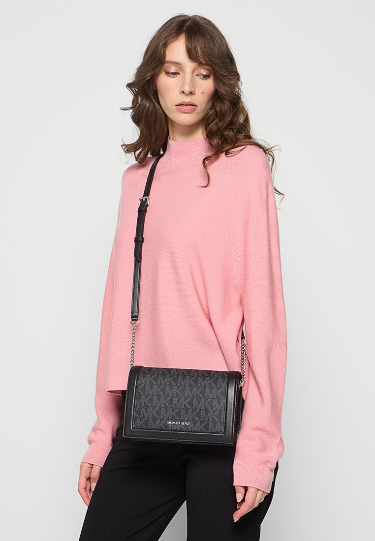 Sacoche en bandoulière noire à motif, avec une texture lisse, une chaîne en tant que bandoulière et un détail de logo. Portée avec un pull rose et un pantalon noir.