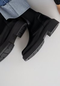 Černé kožené kotníkové boty s textured gumovou podrážkou, masivním podpatkem a hladkým svrškem. Design neobsahuje žádné viditelné zapínání.