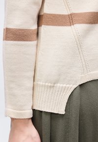 Κοντινή λήψη μανικιού μπεζ πλεκτού πουλόβερ με καφέ ρίγα πάνω από τον αγκώνα, ριμπ μανσέτα και πλαϊνή σχισμή πάνω από ελιά-πράσινο παντελόνι.