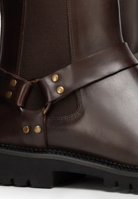 Bottes en cuir marron avec panneaux latéraux élastiques, dotées d'une sangle décorative en cuir et de clous en laiton, montées sur une semelle noire robuste.