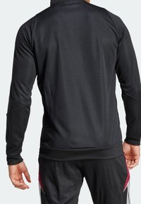 Chaqueta atlética negra de manga larga con una textura suave. Presenta un collar alto y puños ajustados. Combinada con pantalones negros a juego.