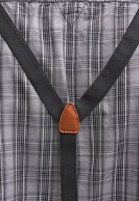 Bretelles noires avec un connecteur en cuir marron portant l'inscription "GUESS" sur un fond en tissu à carreaux gris et blanc.