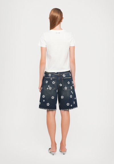 Witte t-shirt met korte mouwen gecombineerd met donkere denimshorts met bloemembroiderie, ruwe zoom en twee achterzakken. Zilveren accenten op de schoenen.