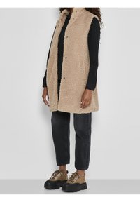 Béžová, načechraná vesta s límečkem a druky, nošená přes černý top. Kombinována s tmavými džínami a robustními hnědými šněrovacími botami.