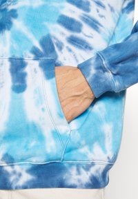 Mano nella tasca di una felpa blu e bianca in tie-dye con polsini e vita a coste, dettagli di cucitura visibili sul tessuto.