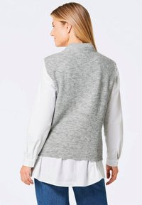 Gilet en laine tricoté gris avec un col montant, fabriqué en laine texturée. Porté sur une chemise blanche, il présente une coupe droite avec une finition lisse.