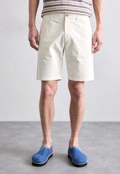 Shorts din bumbac de culoare crem, cu o croială clasică, având închidere cu buton în față și buzunare pe laterale. Purtați cu pantofi slip-on de culoare albastră.