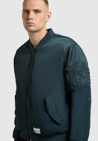 Tmavě zelená bomber bunda s předním zipem, žebrovaným límcem, bočními kapsami a zipovou kapsou na rukávu, s hladkou, lesklou texturou.