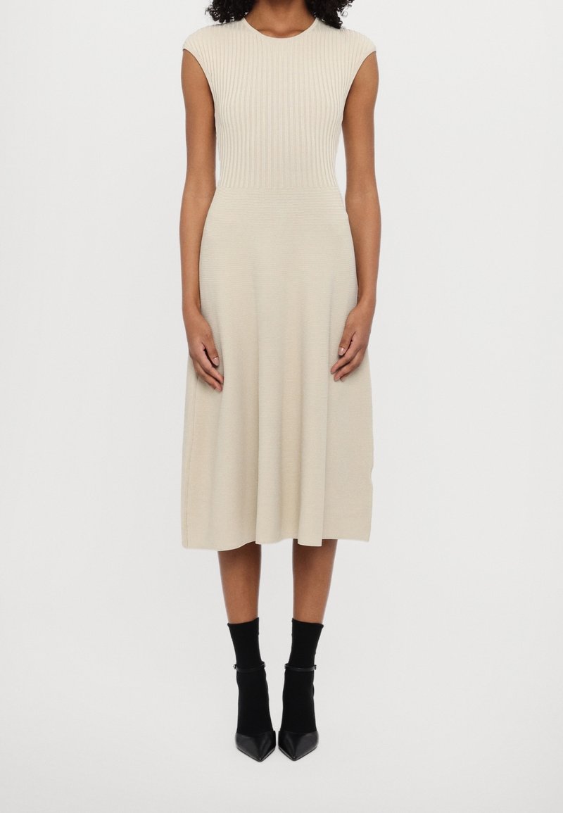 Femme portant une robe midi beige en maille côtelée sans manches avec un corsage ajusté et une jupe évasée, associée à des socquettes noires et des talons pointus.