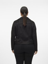 Femme avec une longue queue de cheval noire portant une veste noire et un jean noir, debout de dos sur un fond blanc.